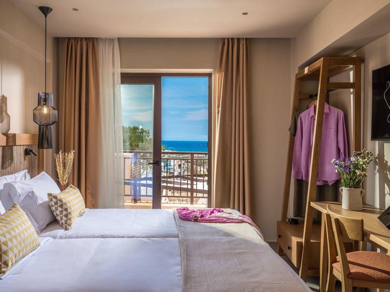Gemütliches Hotelzimmer mit Doppelbett, Balkon und Blick auf das Meer. Helle Holzmöbel, warme Farben.