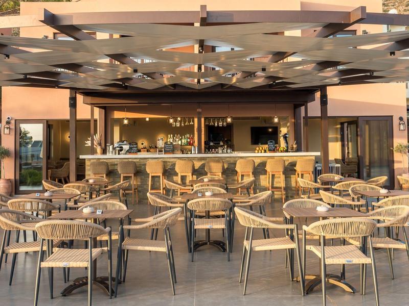 Leerer Außenbereich einer Bar mit Holzstühlen, runden Tischen und großer Pergola bei Sonnenuntergang.