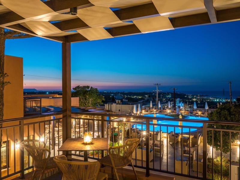 Dachterrasse mit Holztischen und Stühlen, Blick auf beleuchteten Pool und Sonnenuntergang am Meer.