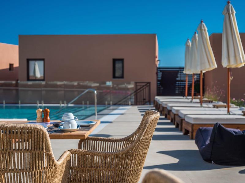 Terrasse mit Pool, Sonnenliegen, Sonnenschirmen und Korbsesseln vor rosa Gebäude.