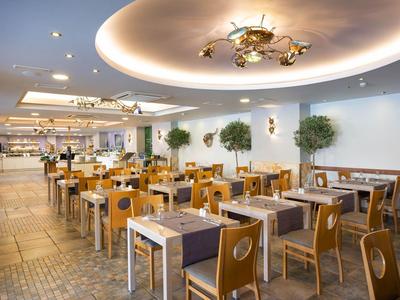 Moderne Restaurant mit Holztischen, Stühlen, weißen Wänden, Pflanzen und futuristischer Deckenbeleuchtung.