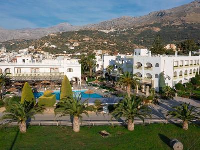 Hotelkomplex mit weißen Gebäuden, Pool, Palmen und Bergen im Hintergrund bei klarem Himmel.