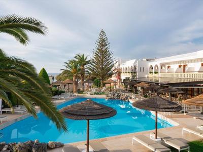 Hotelpool mit blauen Wasser, Sonnenschirmen, Palmen und weißem mehrstöckigem Gebäude im Hintergrund.