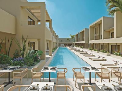 Nowoczesny basen hotelowy z leżakami i stołami po obu stronach, otoczony dwoma skrzydłami budynku.