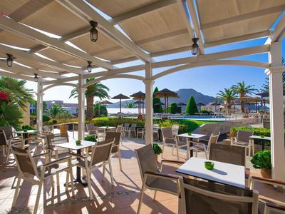 Overdekt terras met tafels en stoelen, uitzicht op zwembad en palmbomen bij zonlicht.