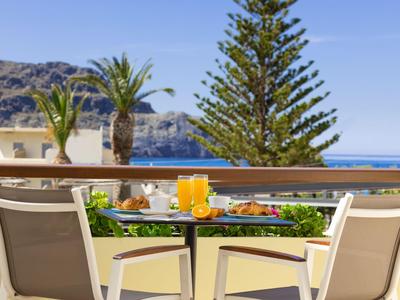 Taras z stołem śniadaniowym, krzesłami i widokiem na palmy i morze w słoneczną pogodę.