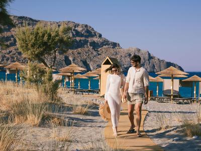 Pareja camina descalza por un sendero de madera hacia la playa con sombrillas y colinas al fondo.