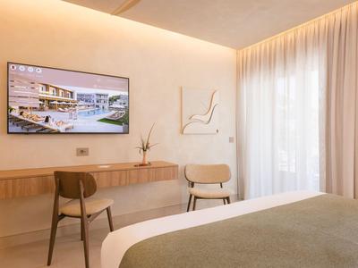 Habitación de hotel luminosa con TV en la pared, dos sillas y gran ventana con cortinas.