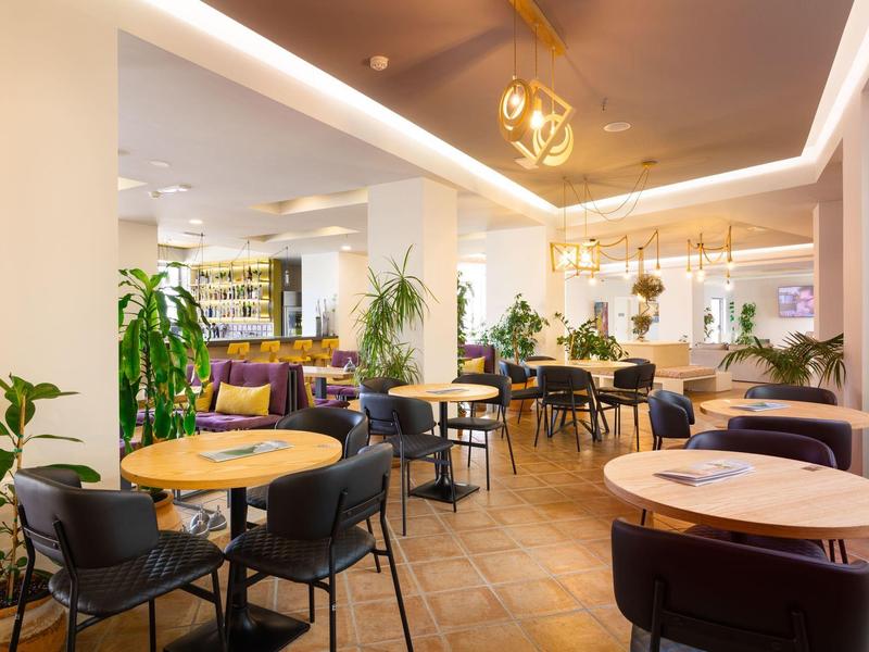 Lichte, moderne hotelbar met ronde houten tafels, zwarte stoelen, planten en stijlvolle verlichting.
