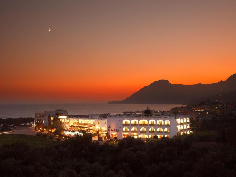 Hotel op kustheuvel met verlichte ramen bij zonsondergang en oranje-rode lucht.