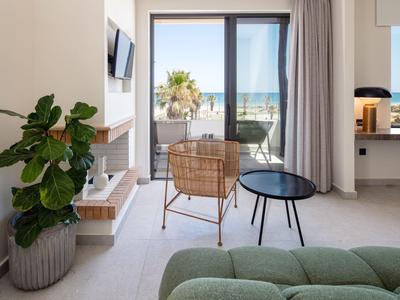 Helles Wohnzimmer mit grüner Couch, Pflanzen, Rattanstuhl und Meerblick durch große Glasfenster.