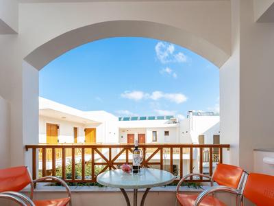 Balkon mit rundem Glas-Tisch und vier roten Stühlen, Blick auf weiße Gebäude und blauen Himmel.