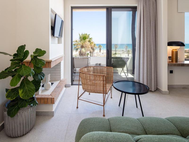 Helles Wohnzimmer mit grüner Couch, Pflanzen, Rattanstuhl und Meerblick durch große Glasfenster.