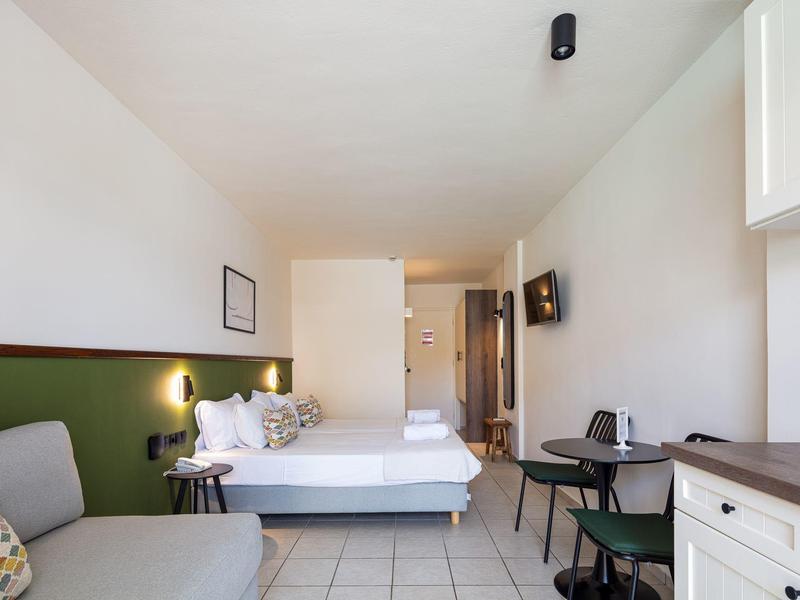 Kleines, helles Zimmer mit Bett, Sofa, Schreibtisch und Fernseher an weiß-grünen Wänden.