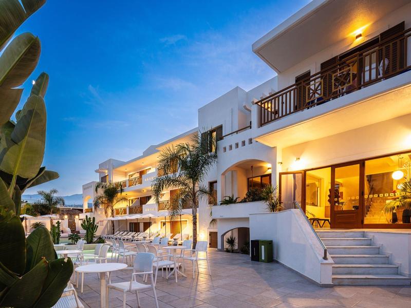 Moderner Hotelhof bei Dämmerung mit weißen Gebäuden, Palmen und Außenmöbeln aus Metall.