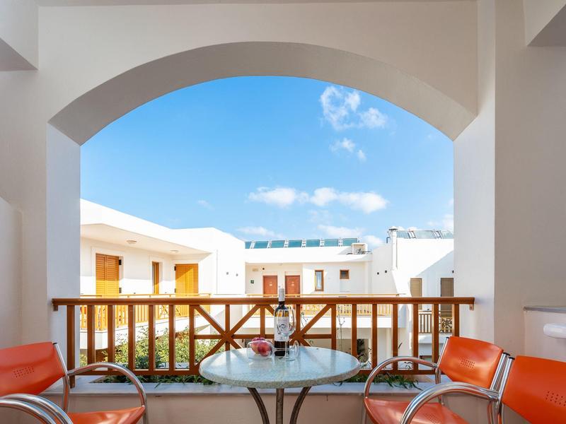 Balkon mit rundem Glas-Tisch und vier roten Stühlen, Blick auf weiße Gebäude und blauen Himmel.