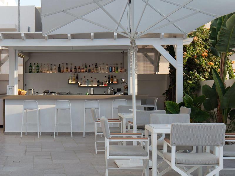 Moderner Außenbereich mit weißem Barwagen, Stühlen und Tischen, umgeben von großen Pflanzen.