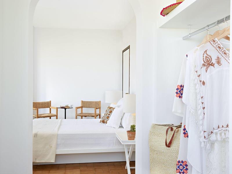 Lichte, eenvoudige hotelkamer met wit beddengoed, twee stoelen en een kleine plant.