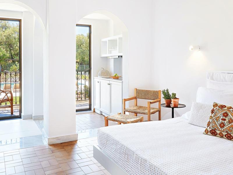 Lichte hotelkamer met tweepersoonsbed, houten stoel, planten en balkon met zeezicht.