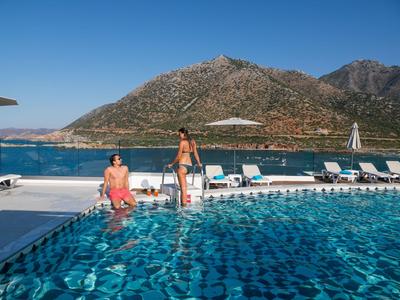 Piscina con sdraio davanti a paesaggio montano e mare, due persone al bordo piscina.