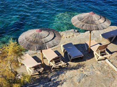 Lettini da spiaggia e ombrelloni di paglia su costa rocciosa con vista sul mare azzurro e limpido.