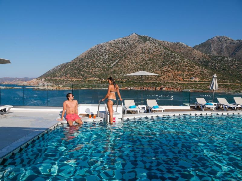 Piscina con sdraio davanti a paesaggio montano e mare, due persone al bordo piscina.