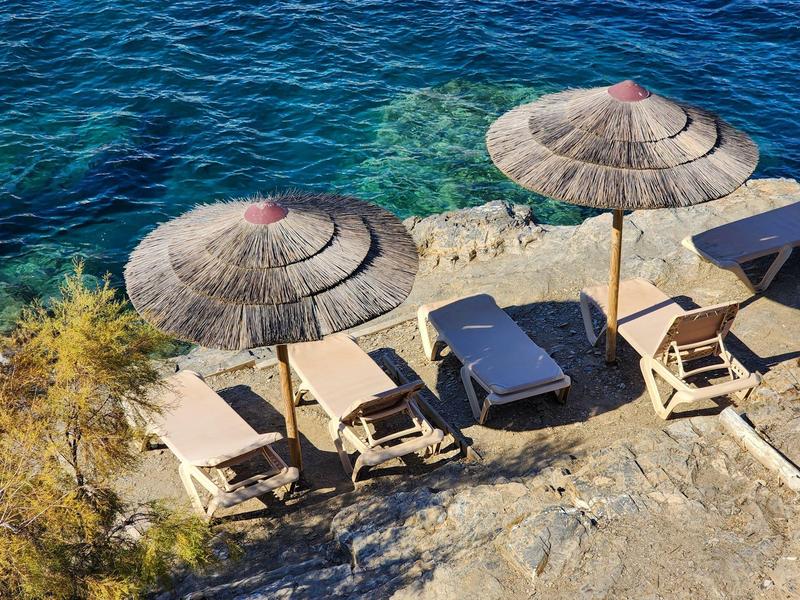 Diverse sdraio con ombrelloni su terreno roccioso accanto ad acqua limpida e blu.