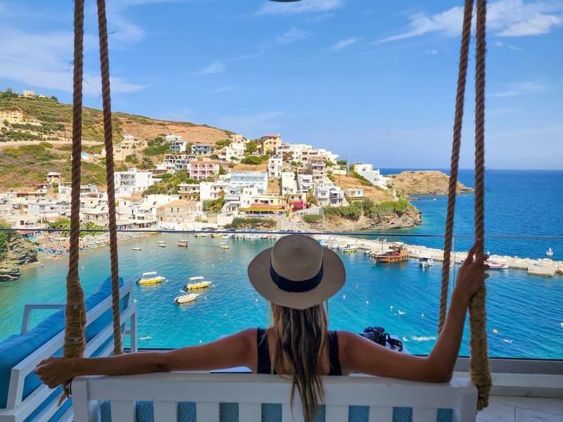 Donna con cappello seduta su un'altalena con vista su una città costiera e il mare blu.
