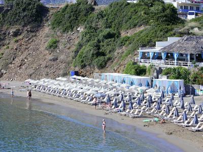 Zandstrand met ligbedden, parasols en gasten bij het water naast een beboste heuvel.