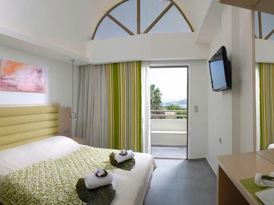 Lichte hotelkamer met tweepersoonsbed, groen-gele gordijnen en balkon met zeezicht.