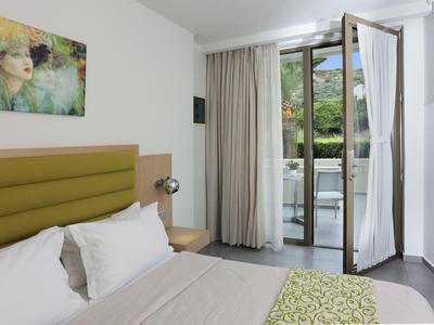 Licht hotelkamer met tweepersoonsbed, open deur en balkon met uitzicht op groene omgeving.