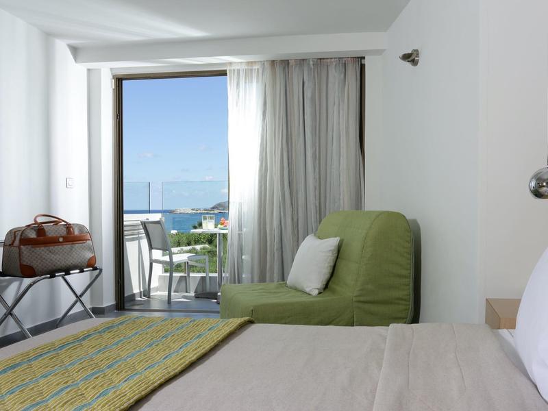 Lichte hotelkamer met bed, groene fauteuil en balkon met uitzicht op zee.