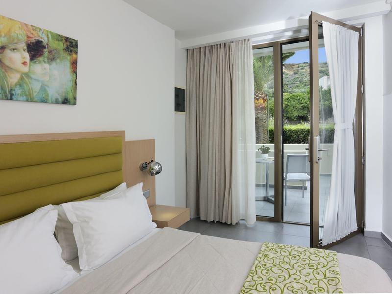 Licht hotelkamer met tweepersoonsbed, open deur en balkon met uitzicht op groene omgeving.