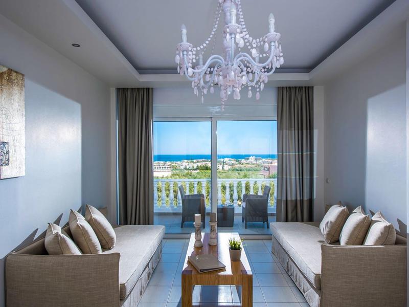 Moderner Wohnraum mit zwei grauen Sofas, Glas-Couchtisch, Kronleuchter und Balkonblick aufs Meer.