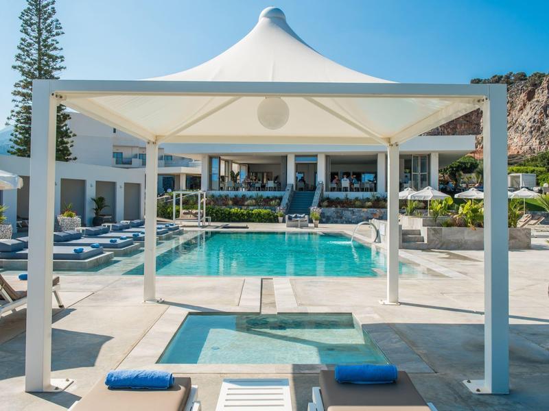 Moderner Außenpool mit überdachter Sitzecke und Liegestühlen in einer Hotelanlage unter blauem Himmel.