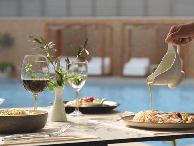 Stůl prostřený s vínem a jídlem u bazénu, ruka nalévá omáčku na jídlo.