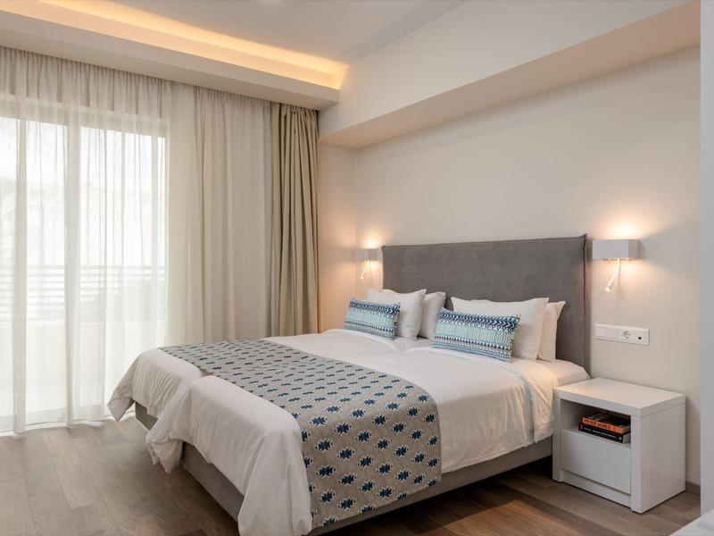 Moderně zařízený hotelový pokoj s manželskou postelí, nočním stolkem a velkými okny se závěsy.