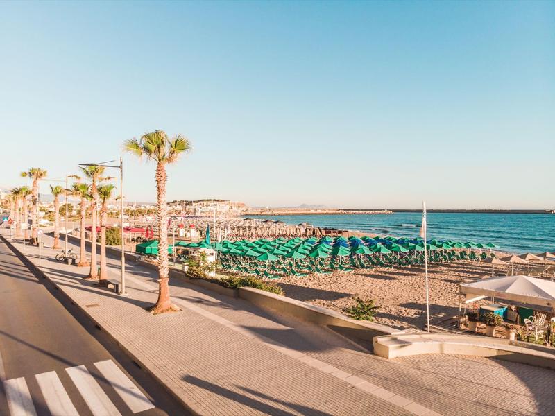 Široká promenáda s palmami vedle pláže se zelenými slunečníky pod modrou oblohou