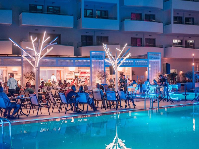 Večerní terasa hotelu s osvětleným bazénem a hosty u stolů.