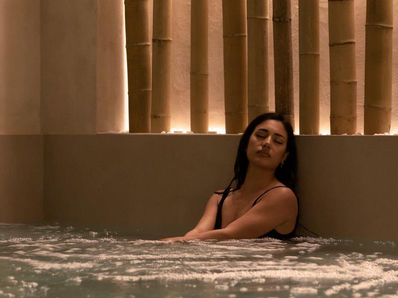 Žena relaxuje v krytém bazénu před zdí s osvětleným bambusem.