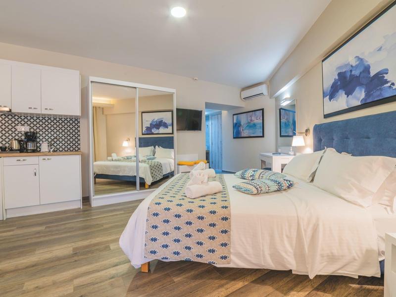 Moderní hotelový pokoj s manželskou postelí, zrcadlovou skříní a kuchyňským koutem.