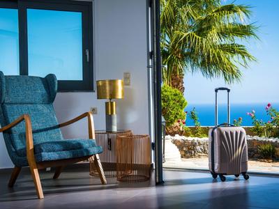 Ein helles Wohnzimmer mit blauem Sessel, goldener Lampe und Koffer vor Meer und Palmen.