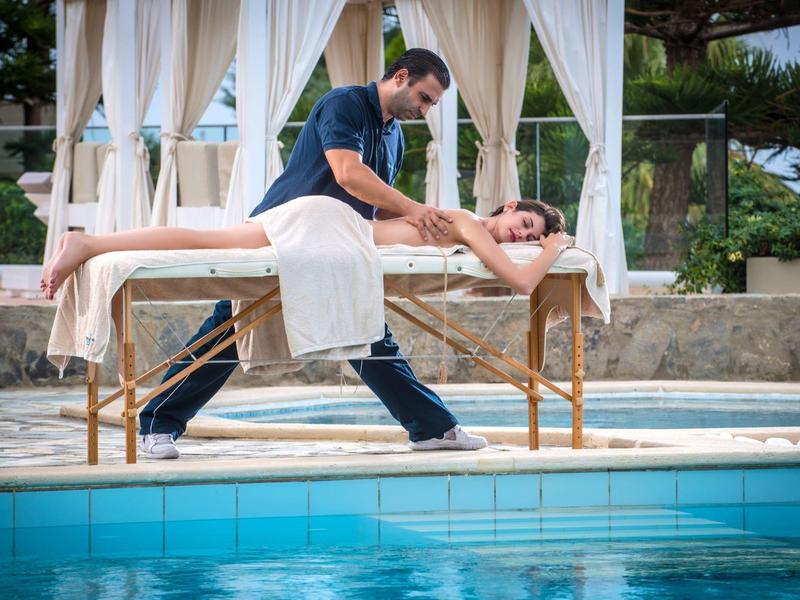 Man massages a woman on a table by a pool under a canopy.