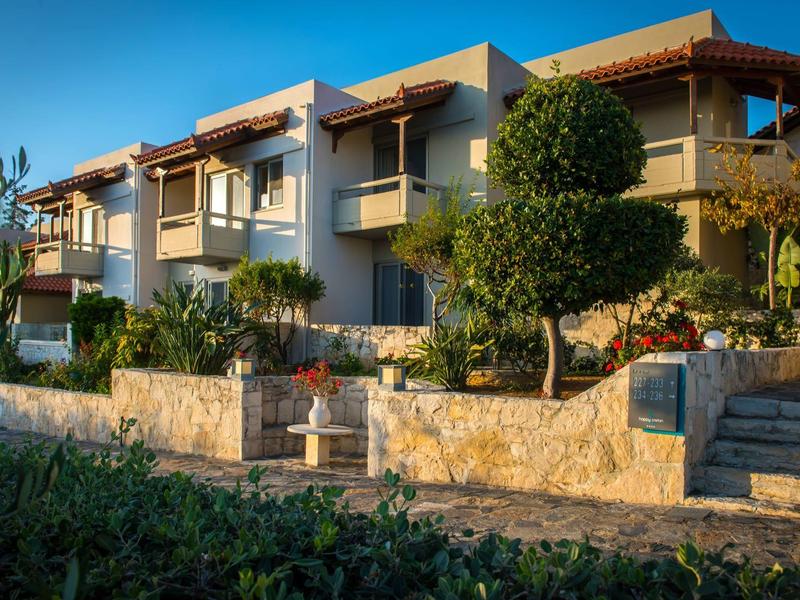 Modern hotel complex with balconies, surrounded by greenery and stone walls at sunset.