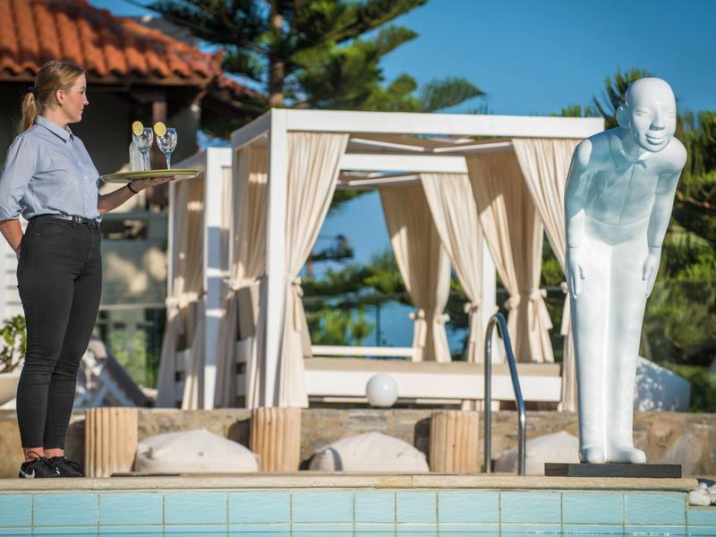 Woman by pool with white sculpture next to a covered lounge in garden.