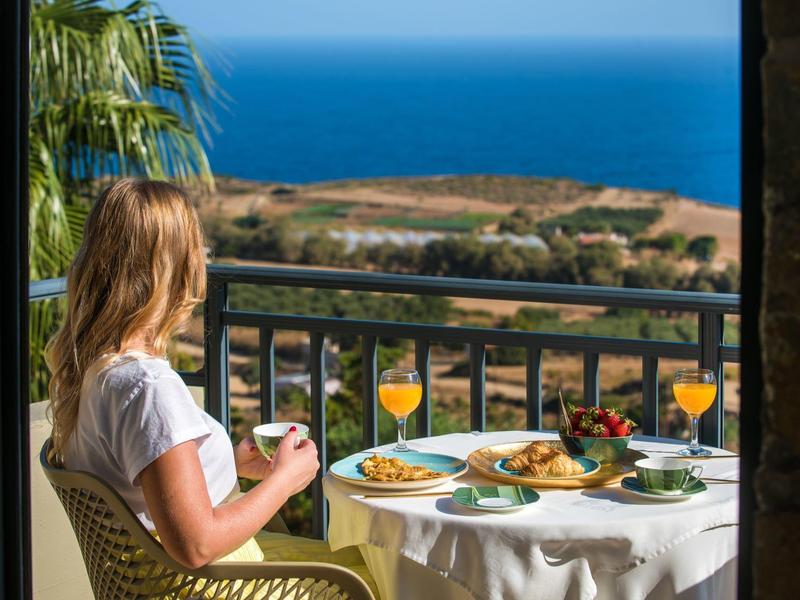 Woman sits on balcony overlooking the sea, eating breakfast with orange juice.