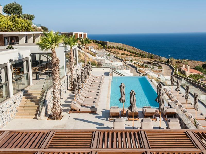 Hotel rooftop terrace with pool, sun loungers, palm trees, and sea view in the background.
