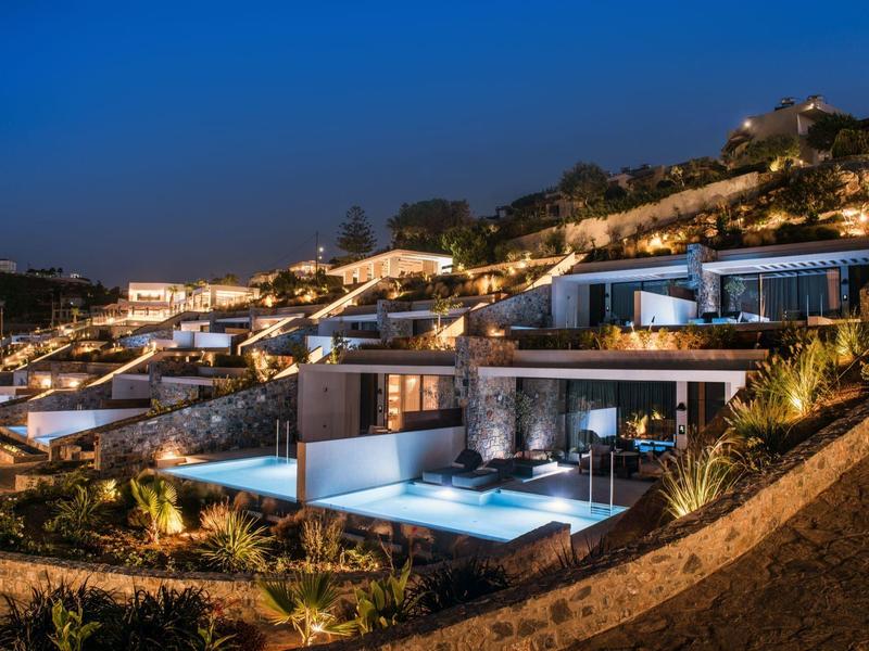 Modern villas with illuminated pools on a hillside under a blue evening sky.