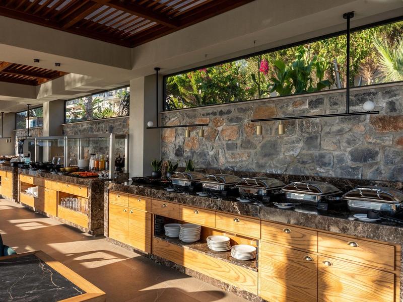 Buffet area with wooden counter, hot food containers, dishes, and large windows overlooking greenery.