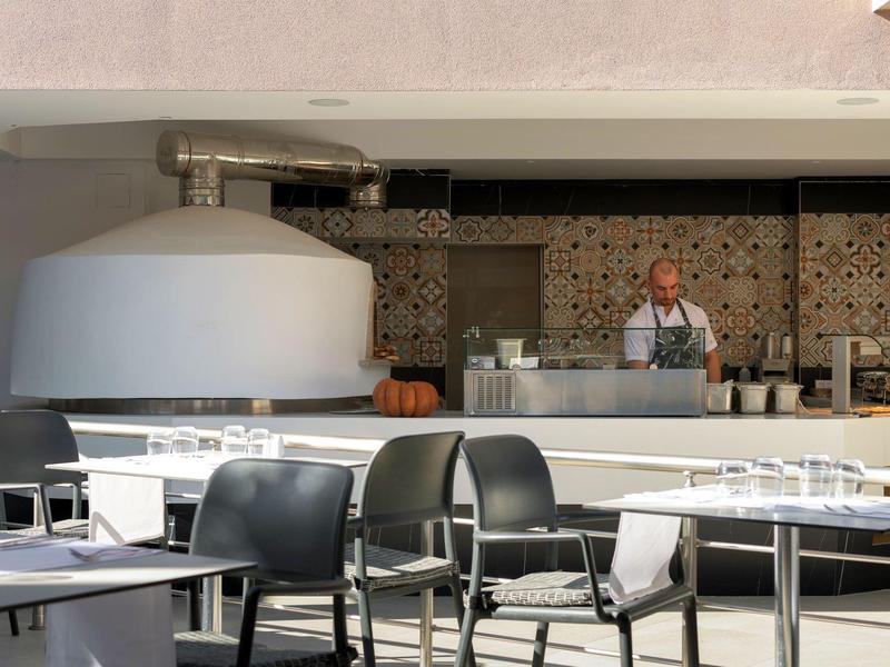 Zewnętrzny obszar restauracyjny z nowoczesnymi czarnymi krzesłami i stołami na słońcu, osoba za barem.